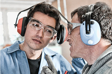 Two workers in a factory wearing safety earmuffs, leaning in to talk to each other amidst loud machinery.