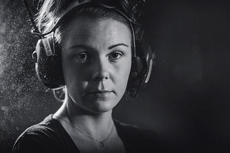 Black and white photo of woman in a factory wearing safety earmuffs,looking at the camera.