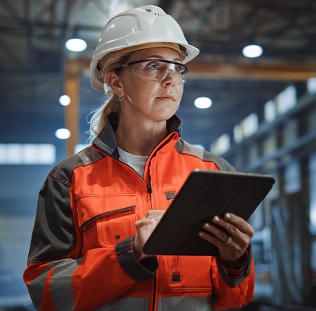 一位身穿高可视性工作服、戴着安全头盔的女性在工厂里手持iPad。